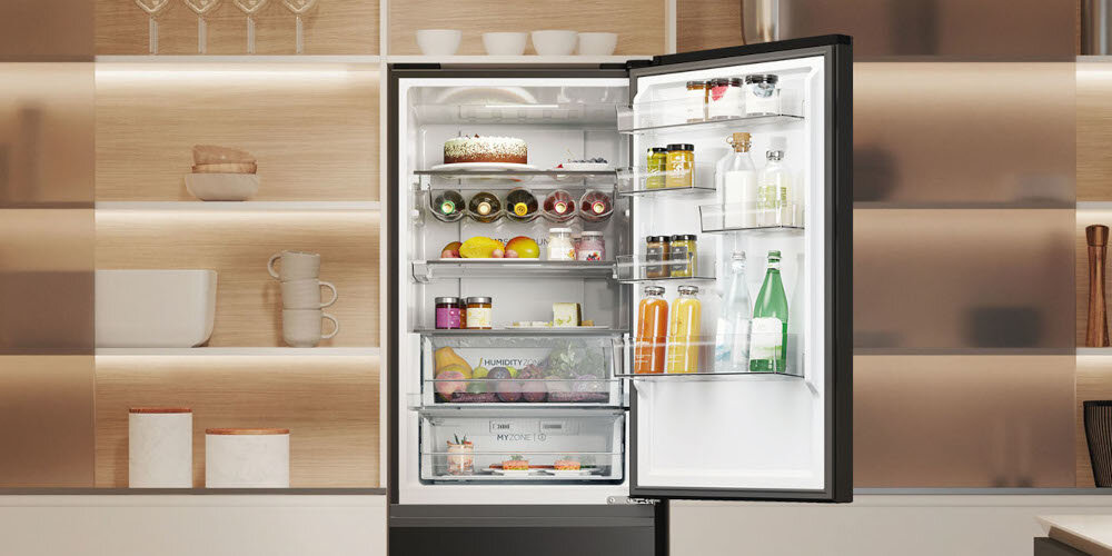 Otwarta czarna lodówka z widocznymi półkami wypełnionymi różnymi produktami spożywczymi, takimi jak owoce, napoje i słoiki. W tle znajduje się nowoczesna kuchnia z drewnianymi i szklanymi elementami.