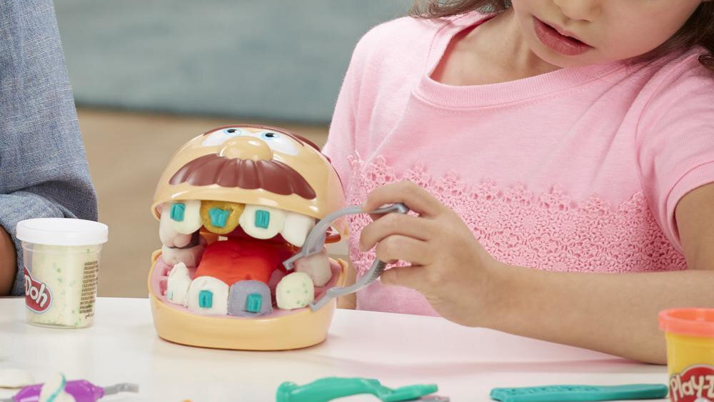 Dziecko bawi się zestawem Ciastolina PLAY-DOH Dentysta, używając narzędzi do modelowania zębów w plastikowej głowie. Obok znajduje się pojemnik z ciastoliną z widocznym logo Play-Doh.