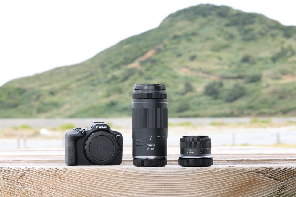 Na drewnianym stole znajdują się trzy elementy fotograficzne: aparat Canon EOS oraz dwa obiektywy, w tym jeden oznaczony jako Canon 75-300 mm. W tle widoczny jest zielony krajobraz z pagórkami.