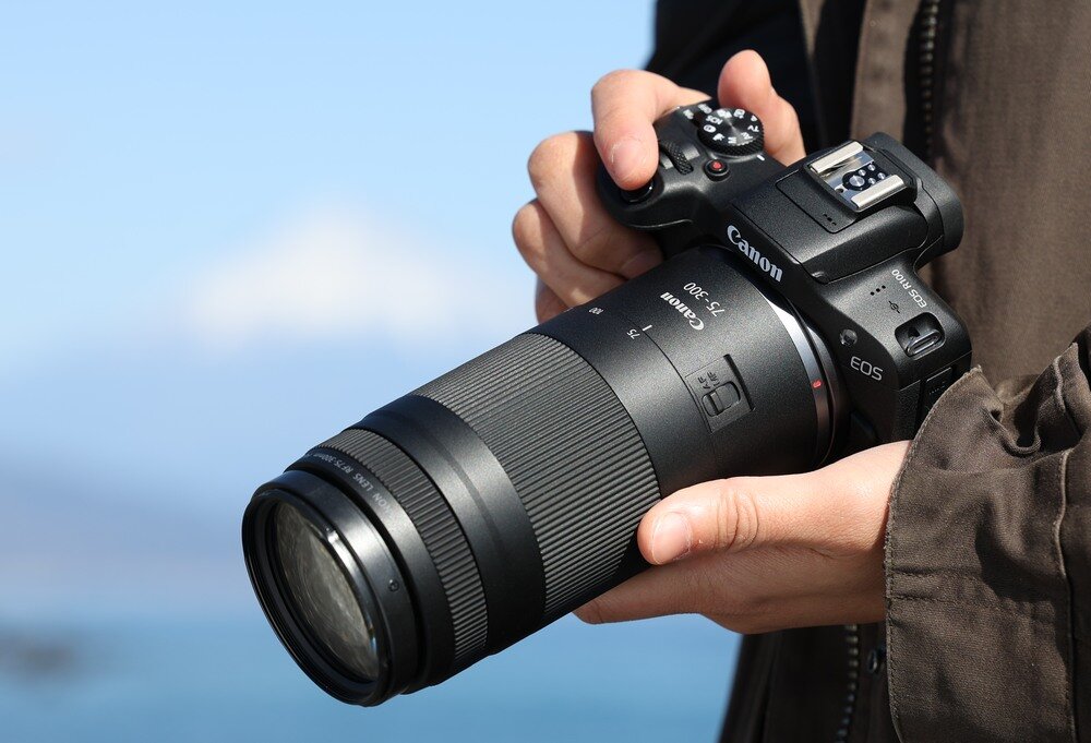 Osoba trzyma aparat fotograficzny z obiektywem Canon RF 75-300 mm f/4-5.6. Na aparacie widoczne jest logo \'Canon\' oraz oznaczenie \'EOS\'.
