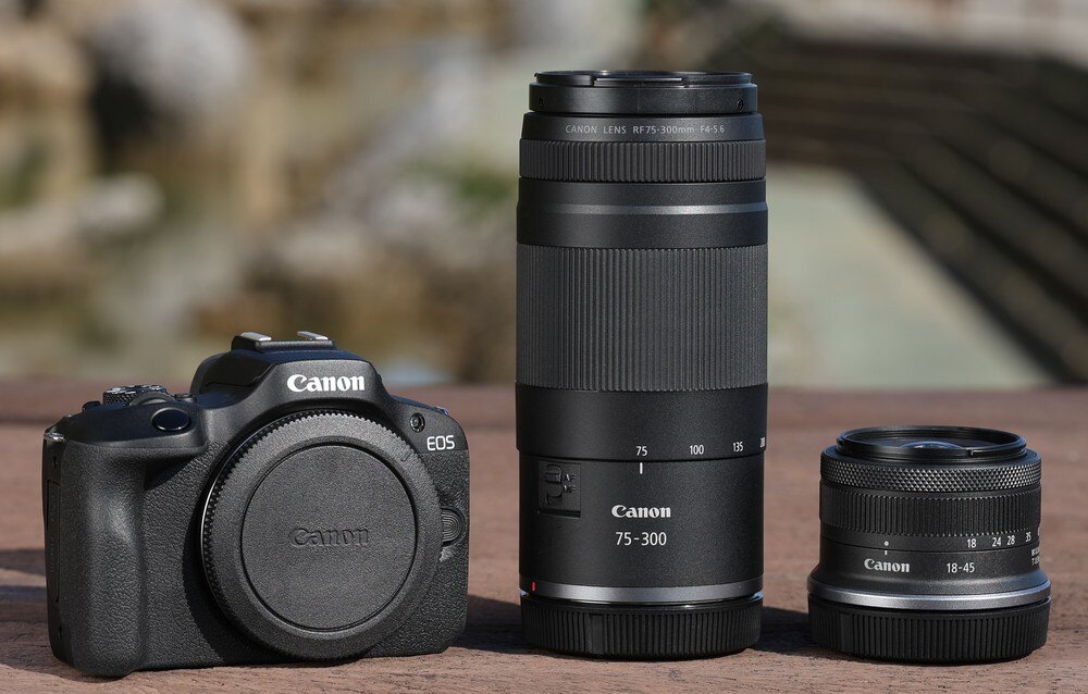 Na drewnianej powierzchni znajdują się trzy elementy fotograficzne: aparat Canon z założonym dekielkiem, obiektyw Canon RF 75-300 mm oraz mniejszy obiektyw Canon 18-45 mm. Wszystkie produkty mają widoczne logo Canon.