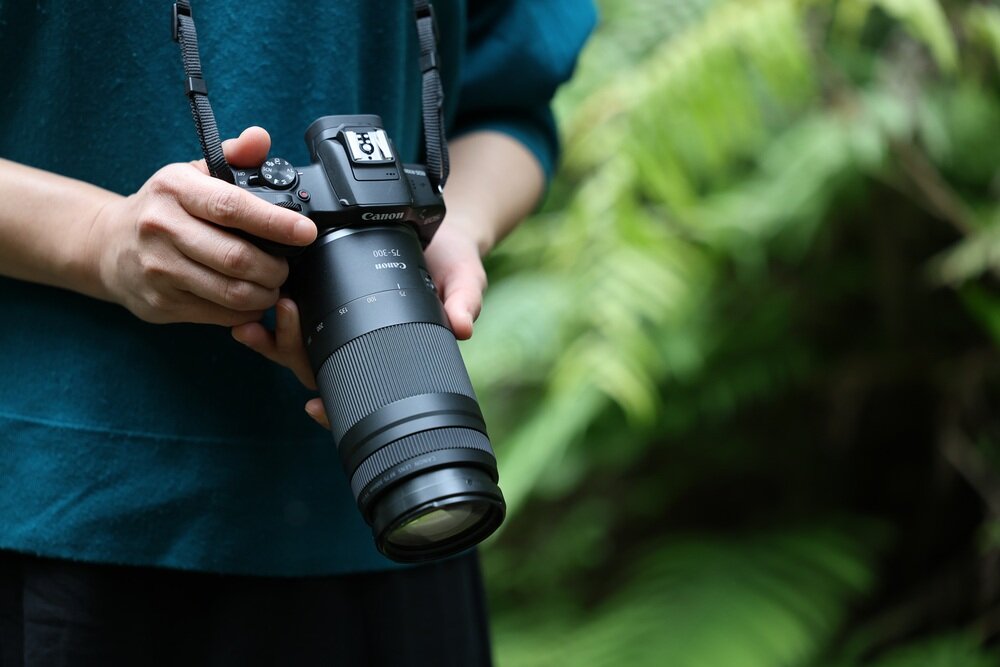 Osoba trzyma aparat z obiektywem Canon RF 75-300 mm f/4-5.6, w tle widoczne są zielone liście. Na aparacie widoczne jest logo Canon.