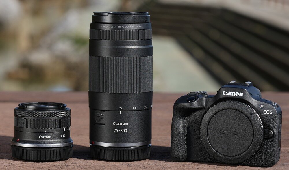 Na drewnianej powierzchni znajdują się dwa obiektywy i korpus aparatu marki Canon. Obiektyw pośrodku ma oznaczenie \'Canon 75-300\'.
