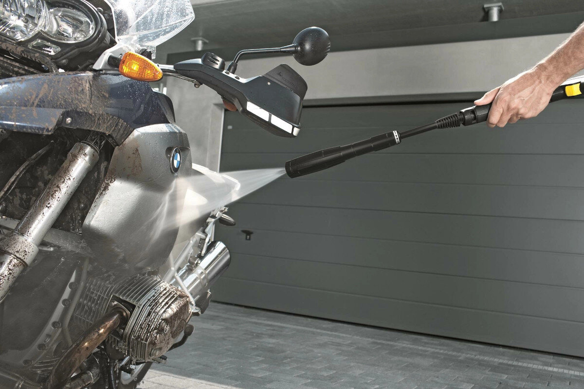Osoba używa myjki ciśnieniowej do czyszczenia motocykla, z którego spływa brud. W tle widoczna jest szara brama garażowa.