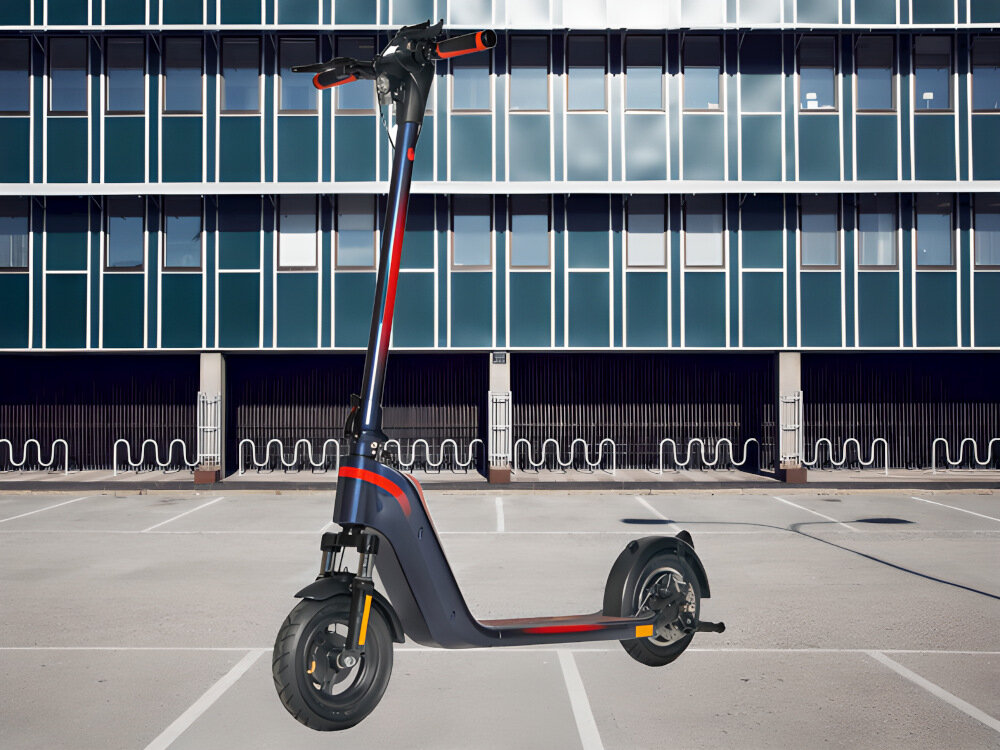 Hulajnoga elektryczna w kolorze granatowo-czerwonym stoi na pustym parkingu przed budynkiem z dużymi oknami. Hulajnoga ma duże koła i prostą kierownicę.
