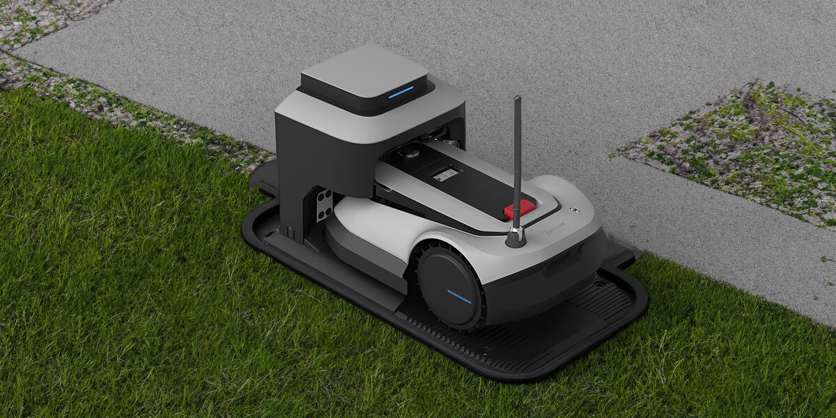 Na obrazie widoczny jest szary robot koszący umieszczony na trawie, z anteną i czarnymi elementami. Robot znajduje się na czarnej podstawce, a w tle widać fragment chodnika.