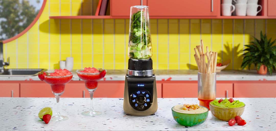 Na blacie kuchennym stoi blender z logo Tefal, wypełniony zielonymi składnikami. Obok znajdują się dwa kieliszki z czerwonym napojem, miski z przekąskami oraz pojemnik z paluszkami.