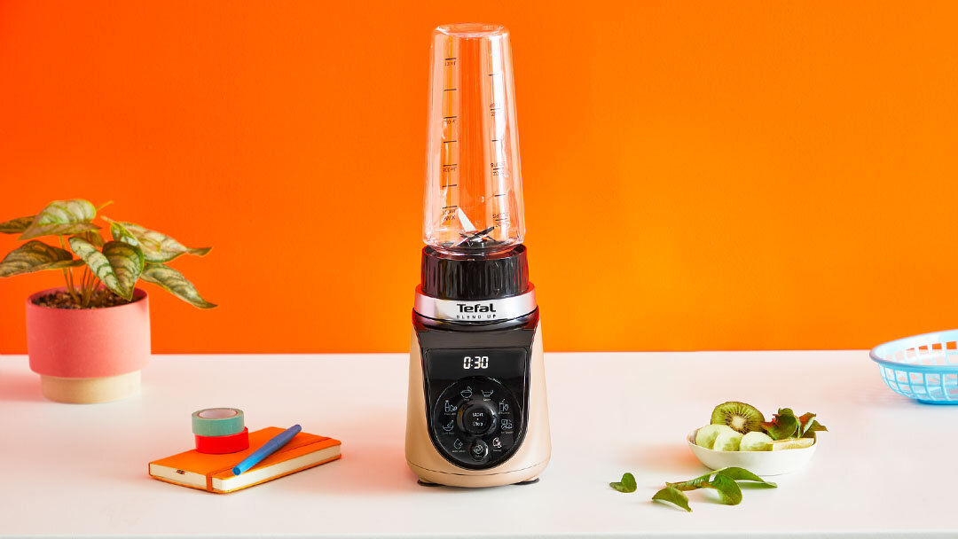 Na białym blacie stoi blender z przezroczystym pojemnikiem i czarnym panelem sterowania z wyświetlaczem pokazującym \'0:30\'. Obok znajdują się rośliny doniczkowe, notes z długopisem oraz miska z pokrojonymi owocami.