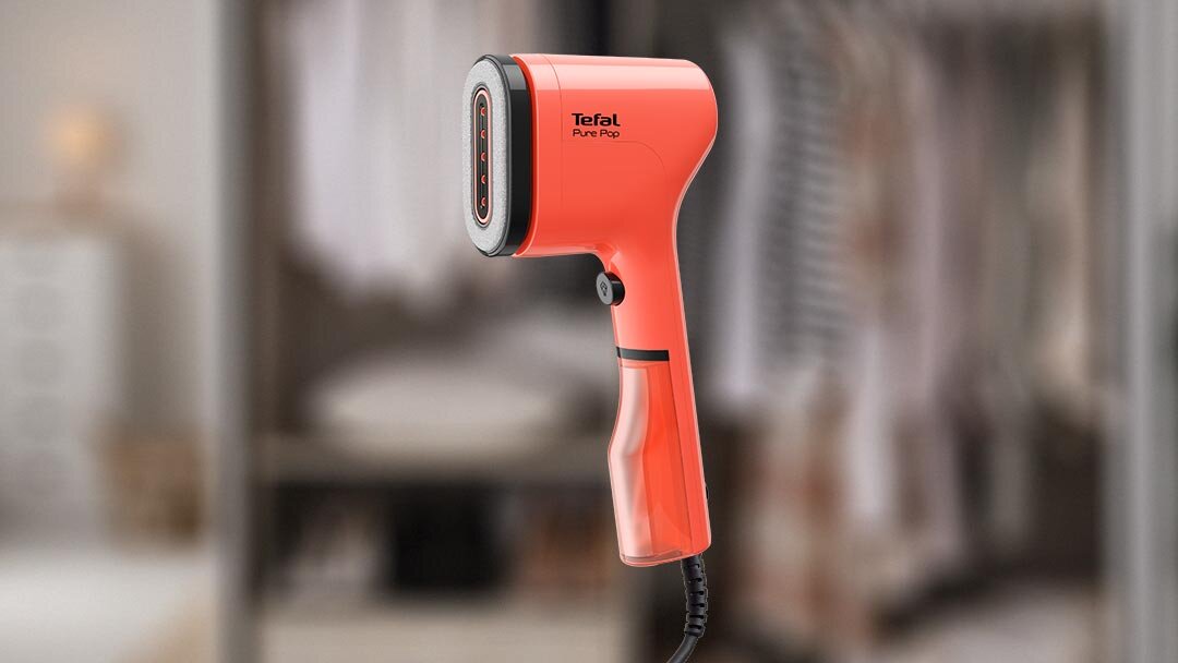 Na obrazie widoczna jest czerwona parownica do ubrań z napisem \'Tefal Pure Pop\'. W tle znajduje się rozmyta szafa z ubraniami.