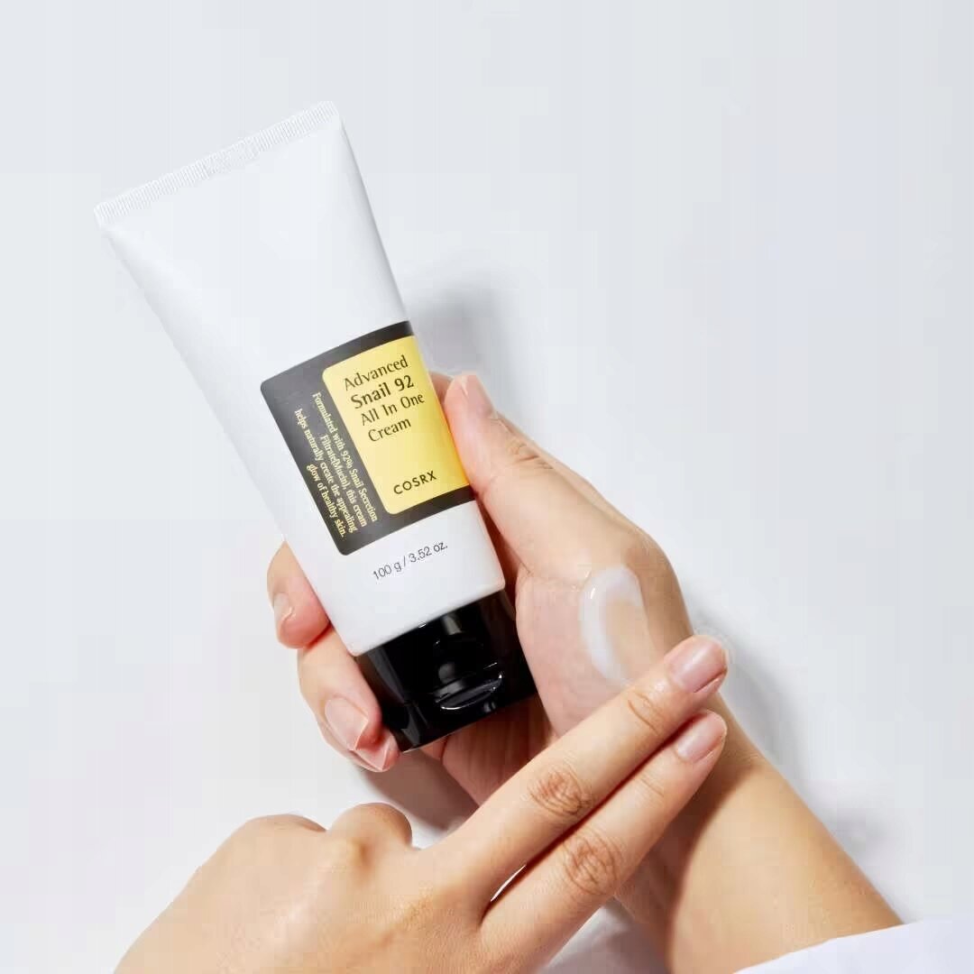 Osoba trzyma tubkę kremu do twarzy z napisem \'Advanced Snail 92 All In One Cream COSRX\'. Na dłoni widoczna jest niewielka ilość kremu.