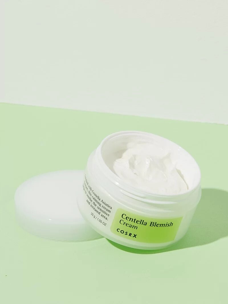 Otwarty słoik z białym kremem na jasnozielonym tle. Na etykiecie widnieje napis: \'Centella Blemish Cream COSRX\'.