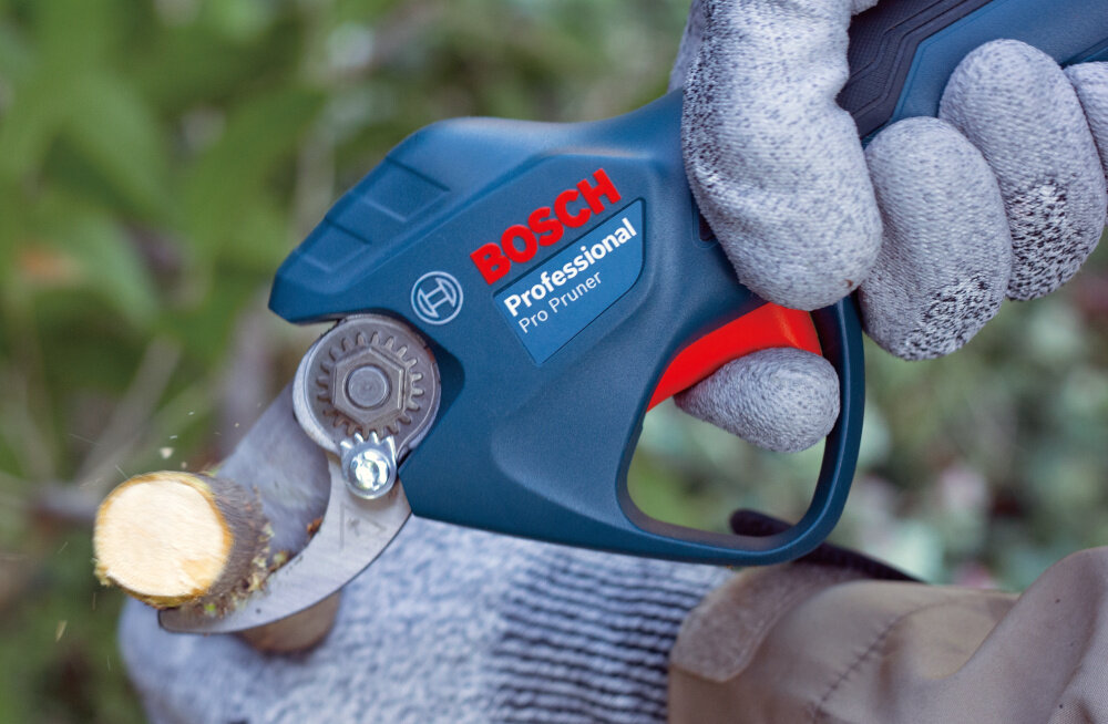 Osoba w rękawicach trzyma sekator do gałęzi z widocznym logo \'BOSCH Professional Pro Pruner\'. Sekator przecina gałąź, a w tle widać rozmytą zieleń.