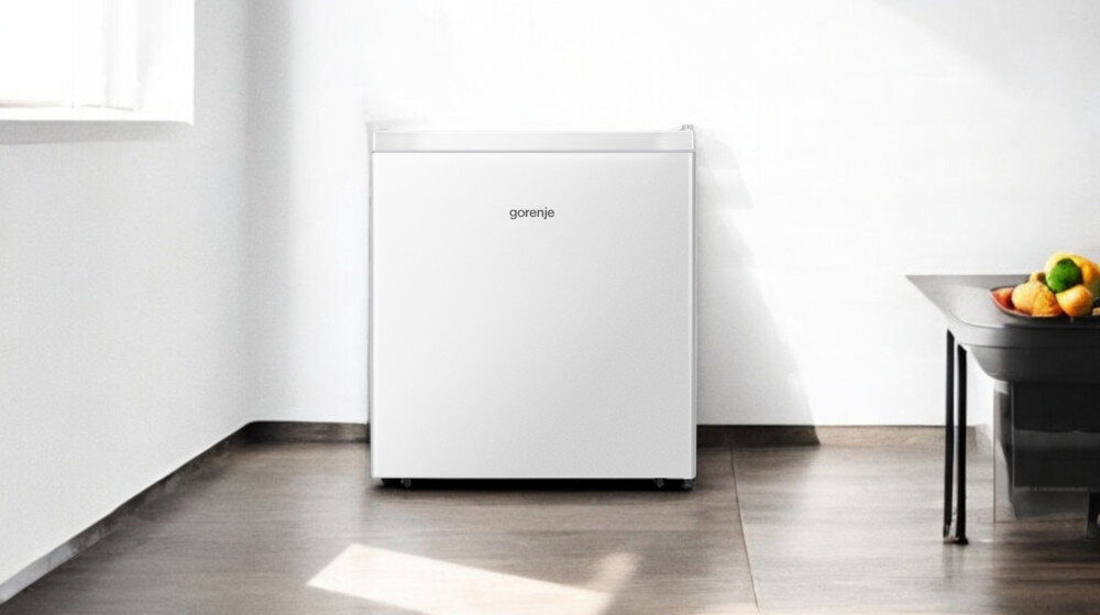 Biała lodówka z widocznym logo \'gorenje\' stoi w jasnym pomieszczeniu na drewnianej podłodze. Obok znajduje się stół z miską pełną owoców.
