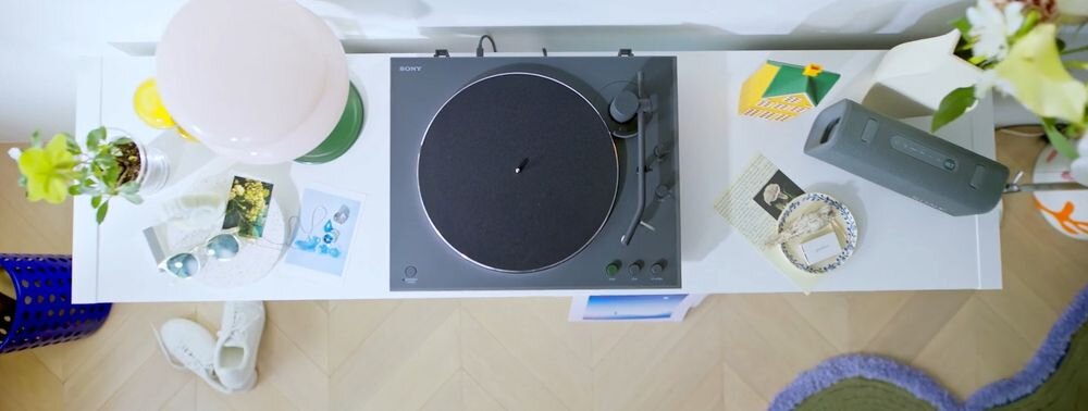 Na białej półce znajduje się czarny gramofon z logo SONY, otoczony różnymi przedmiotami, takimi jak lampa, okulary przeciwsłoneczne i przenośny głośnik. Obok gramofonu leżą zdjęcia, rośliny i inne drobne akcesoria.