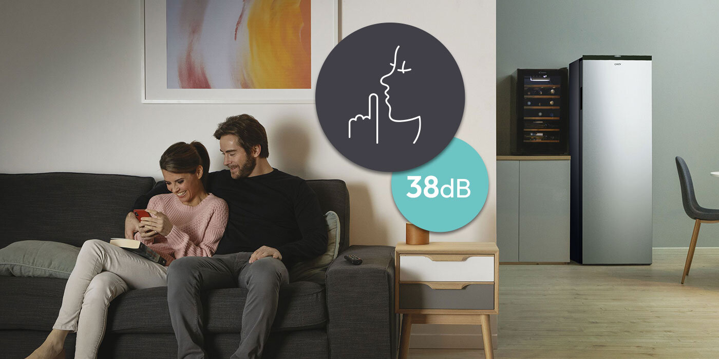 Para siedzi na kanapie w salonie, obok stoi zamrażarka w kolorze srebrnym. Na obrazie widoczna jest ikona z symbolem ciszy i tekstem \'38dB\'.