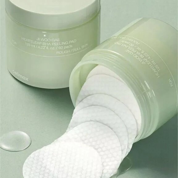 Na obrazie widoczne są dwa otwarte pojemniki z białymi płatkami kosmetycznymi. Na pojemnikach znajduje się tekst: \'JI WOO GAE HEARTLEAF BHA PEELING PAD 125 ml / 4.22 fl. oz. / 60 pads ROUGH / DULL SKIN\' oraz logo \'celimax\'.
