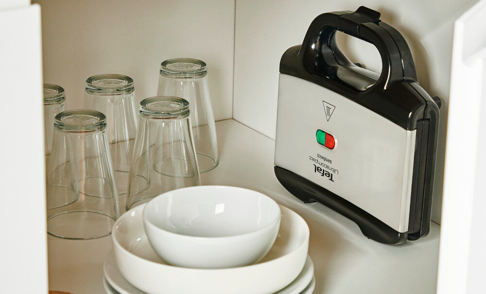 Na zdjęciu widać opiekacz z logo \'Tefal\', stojący na półce obok kilku odwróconych szklanek i białych misek. Opiekacz ma dwa przyciski: zielony i czerwony, oraz symbol trójkąta z wykrzyknikiem.