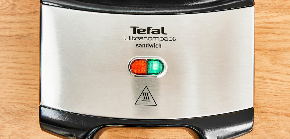 Na obrazie widoczny jest opiekacz z metaliczną obudową i dwoma kontrolkami: czerwoną i zieloną. Na obudowie znajduje się tekst: \'Tefal Ultracompact sandwich\' oraz symbol pary.
