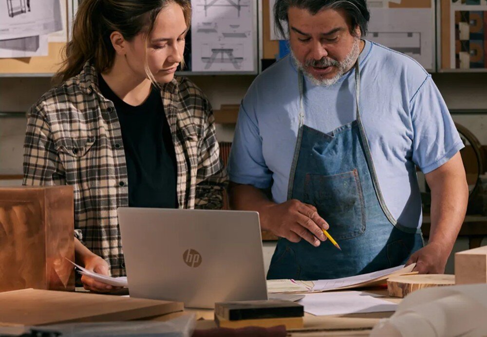 Na stole znajduje się otwarty laptop z widocznym logo HP. W tle widać dwie osoby przeglądające dokumenty oraz różne projekty na ścianie.