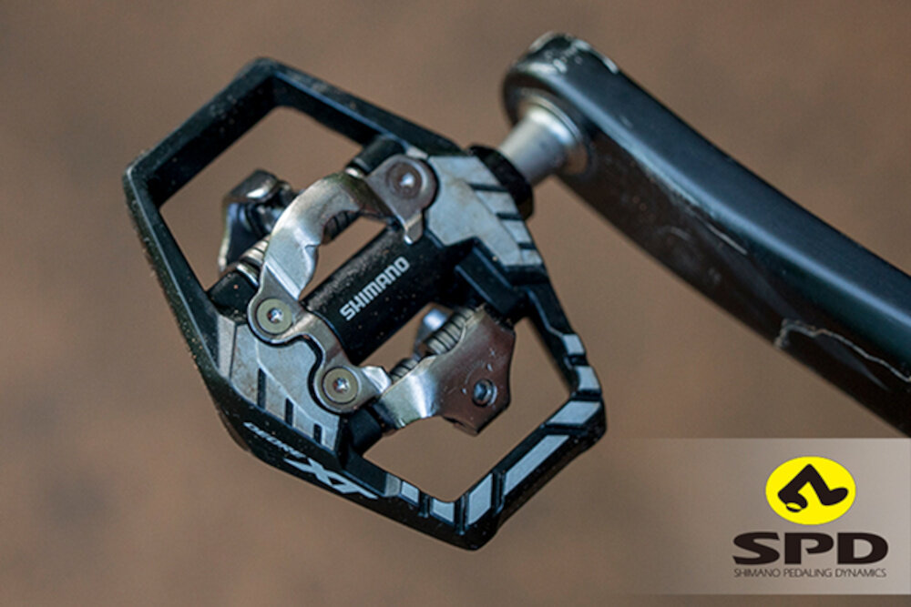 Zdjęcie przedstawia pedał rowerowy z logo SHIMANO. W prawym dolnym rogu widoczne jest logo SPD z napisem \'SHIMANO PEDALING DYNAMICS\'.
