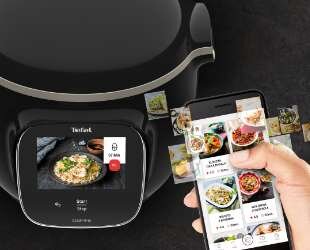 Na obrazie widoczny jest multicooker z wyświetlaczem pokazującym danie oraz aplikacja na smartfonie z przepisami kulinarnymi. Logo \'Tefal\' znajduje się na wyświetlaczu urządzenia.