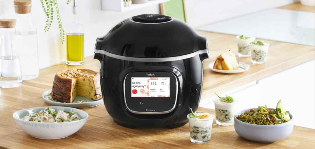 Na blacie kuchennym stoi czarny multicooker z wyświetlaczem, na którym widoczny jest tekst: \'Co dziś ugotujemy?\'. Wokół urządzenia znajdują się różne potrawy w miseczkach i szklankach.