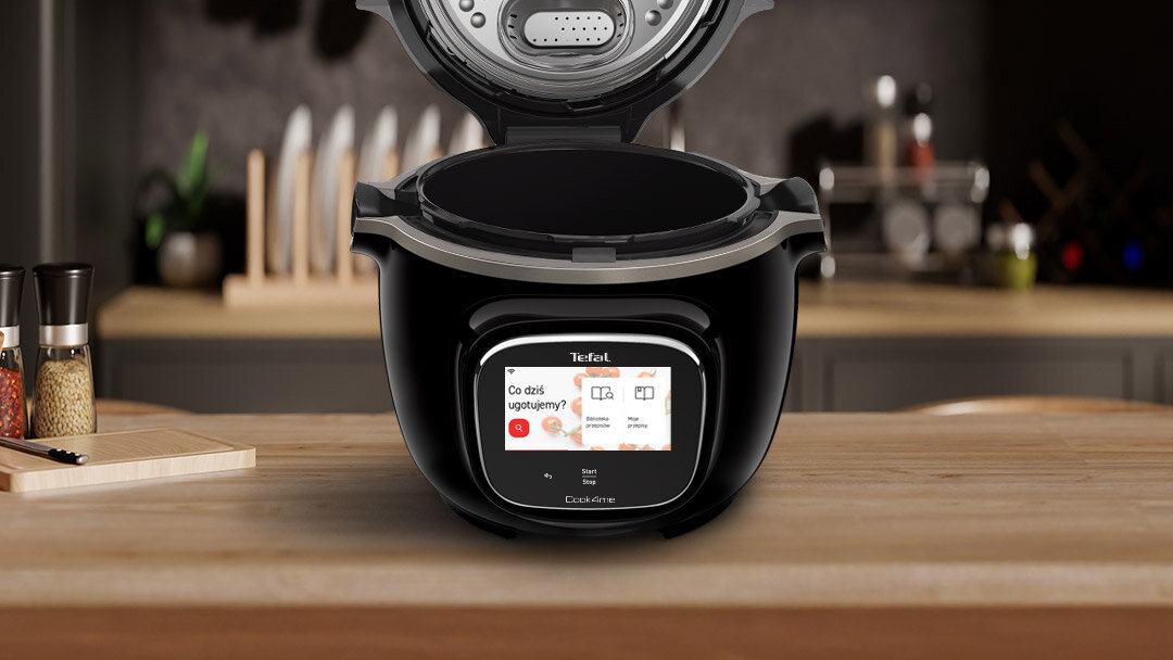 Na blacie kuchennym stoi multicooker z otwartą pokrywą, na wyświetlaczu widoczny jest tekst: \'Co dziś ugotujemy?\'. W tle znajdują się noże i przyprawy.