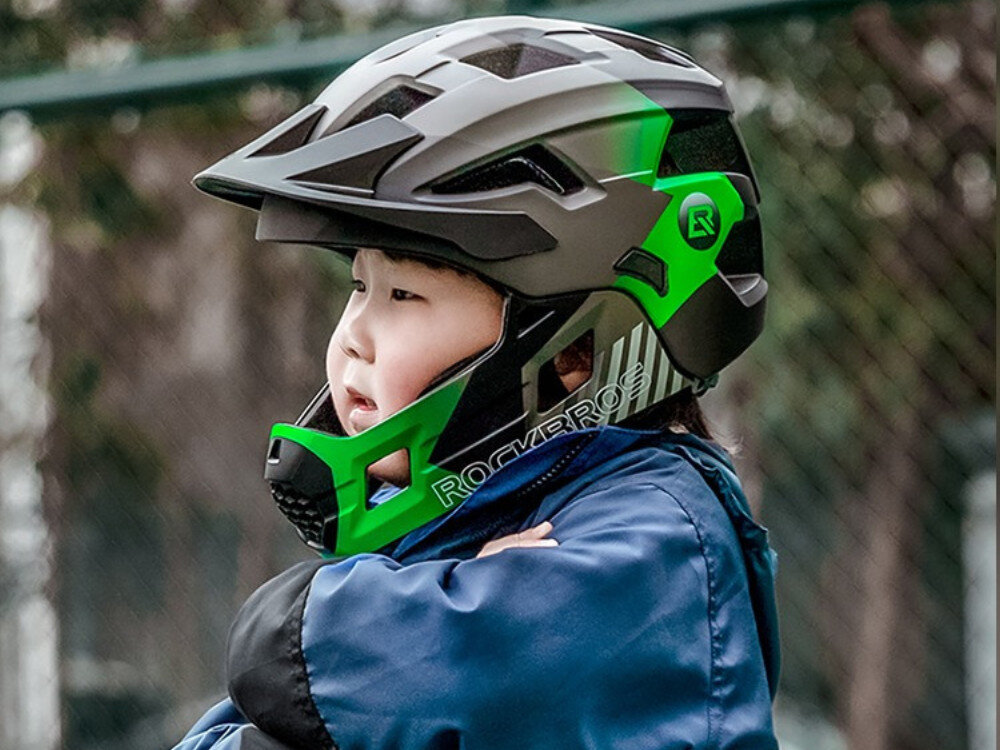 Dziecko nosi zielono-czarny kask rowerowy z odkręcaną szczęką, na którym widoczne jest logo \'ROCKBROS\'.