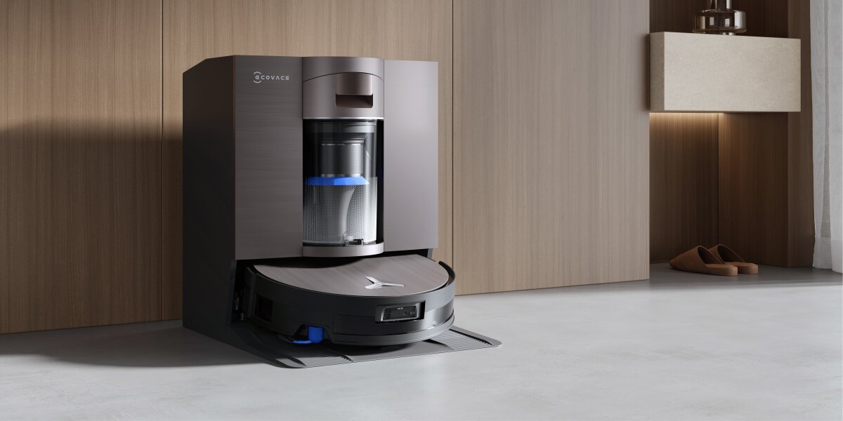 Na obrazie widoczny jest czarny robot sprzątający umieszczony w stacji dokującej z logo \'ECOVACS\'. W tle znajduje się drewniana ściana oraz para butów.
