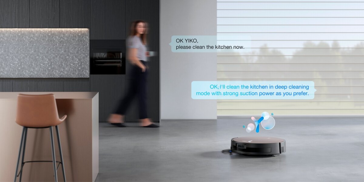 W nowoczesnej kuchni na podłodze znajduje się czarny robot sprzątający. Obok robota widoczne są dwa komunikaty: \'OK YIKO, please clean the kitchen now.\' oraz \'OK, I\'ll clean the kitchen in deep cleaning mode with strong suction power as you prefer.\'