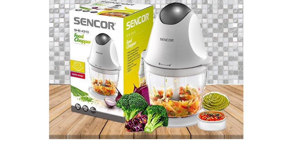 Na obrazie znajduje się rozdrabniacz do warzyw SENCOR SHB 4310 oraz jego opakowanie z widocznym logo SENCOR i napisem \'Food Chopper\'. Przed urządzeniem leżą różne warzywa, w tym brokuł i kapusta, a także miseczki z pokrojonymi warzywami i zielonym sosem.