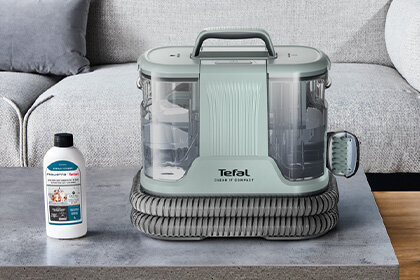 Na szarej powierzchni stoi odkurzacz piorący z napisem \'Tefal Clean It Compact\' oraz butelka środka czyszczącego z widocznym tekstem \'Proszek do prania\'.