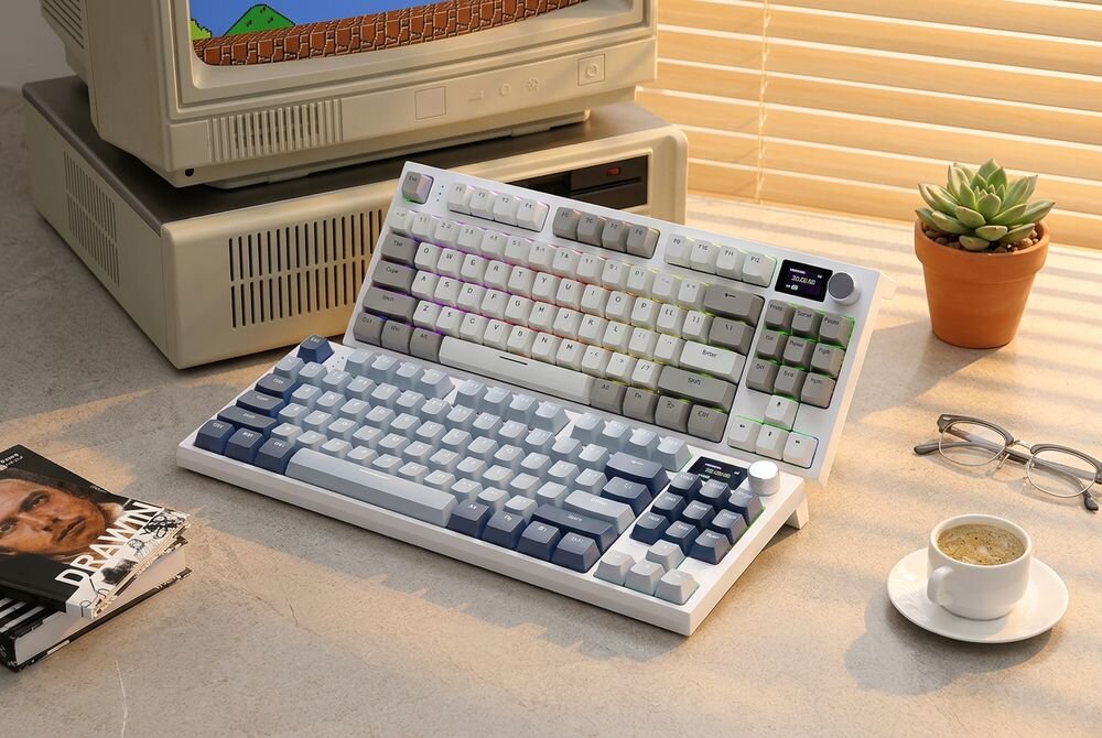 Na biurku znajduje się biało-szara klawiatura z podświetleniem RGB, obok której leży filiżanka kawy, okulary i magazyn. W tle widać stary komputer i doniczkę z sukulentem.