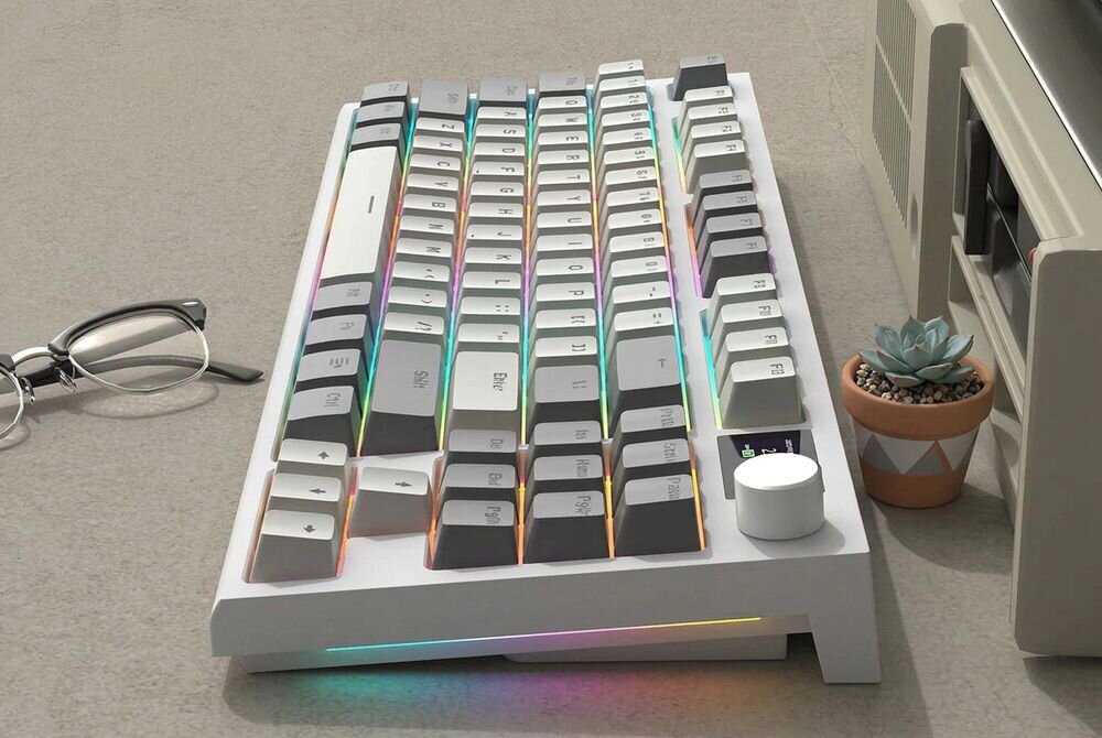 Na obrazie widoczna jest klawiatura w kolorze biało-szarym z podświetleniem RGB. Obok klawiatury znajdują się okulary i mała roślina w doniczce.