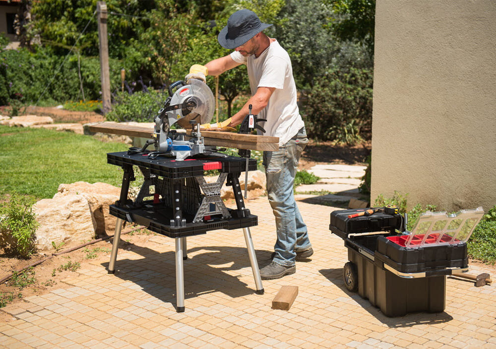 Stół warsztatowy składany KETER Folding Work Table 453 kg podwórko osoba pracuje przy stole obok sto skrzynka narzędziowa w tle drzewa mobilne stanowisko pracy wszechstronne zastosowanie