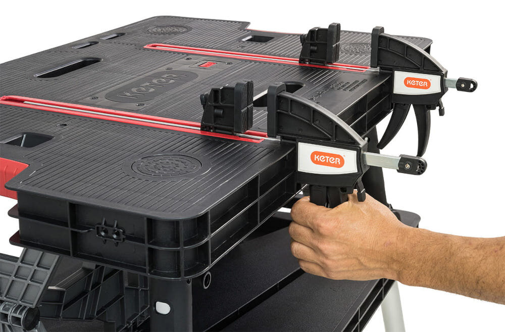 Stół warsztatowy składany KETER Folding Work Table 453 kg zbliżenie na blat osoba trzyma dłoń na jednym z zacisków dwa zaciski o długości roboczej 310 mm pewne unieruchomienie obrabianych elementów materiał pozostaje stabilny precyzja i bezpieczeństwo ogranicza ryzyko błędów większa dokładność wykonania