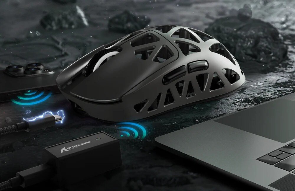 Mysz ATTACK SHARK R2 Ażurowa mysz na stole obok laptopa i telefonu. Pod myszą widać piktogram zasięgu bezprzewodowego, a obok odłączony kabel USB z grafiką błyskawicy