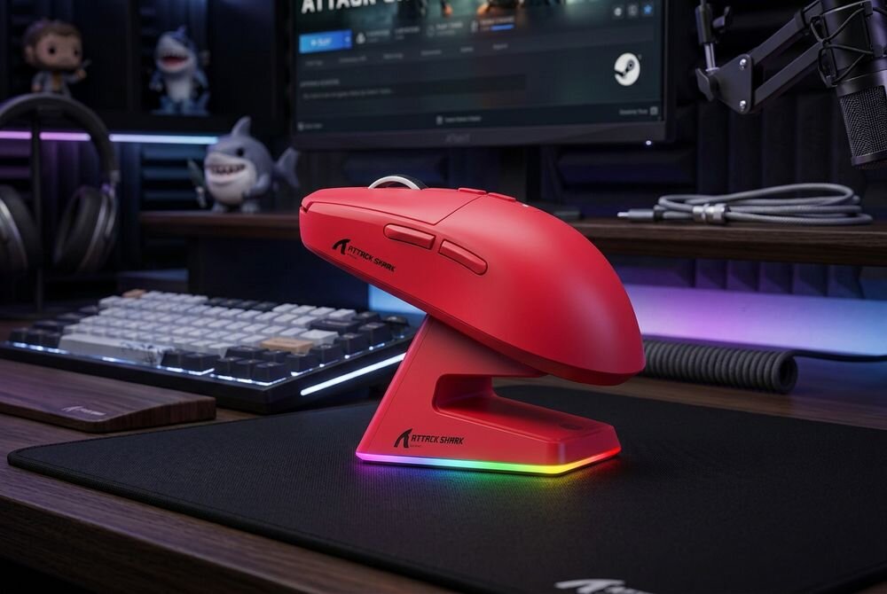 Czerwona mysz komputerowa na podstawce z podświetleniem RGB, z widocznym logo \'ATTACK SHARK\'. W tle znajduje się klawiatura, monitor oraz figurki rekinów.