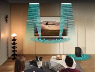 W salonie znajduje się telewizor z wyświetlanym obrazem samolotu, a pod nim soundbar z efektami dźwiękowymi wizualizowanymi jako niebieskie fale. Obok stoi bezprzewodowy subwoofer, a na kanapie siedzą dwie osoby z psem.