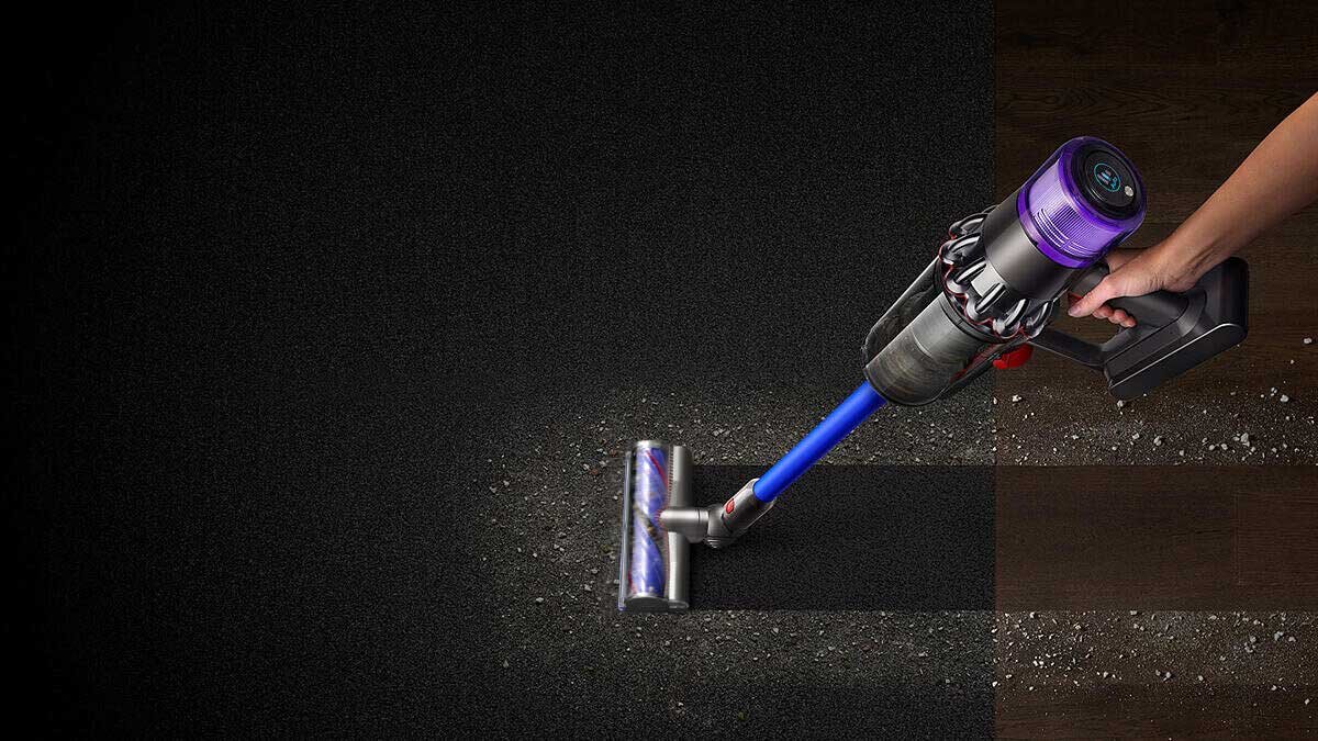 Osoba trzyma odkurzacz z fioletowym i niebieskim akcentem, który zbiera kurz z ciemnej powierzchni. Odkurzacz ma przezroczystą rurę ssącą i widoczny jest wirnik.
