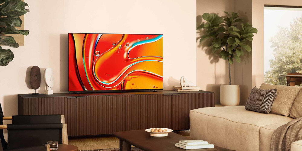 W salonie na szafce stoi duży telewizor z wyświetlonym kolorowym obrazem abstrakcyjnym. Obok znajduje się jasna sofa i roślina w doniczce.