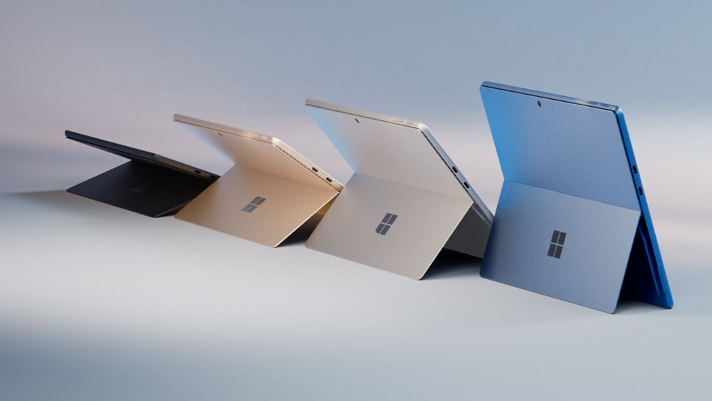 Na obrazie widać cztery laptopy w różnych kolorach, ustawione pod kątem, z widocznym logo w kształcie okna. Laptopy mają rozłożone podpórki, a ich obudowy są w odcieniach czerni, złota, srebra i niebieskiego.