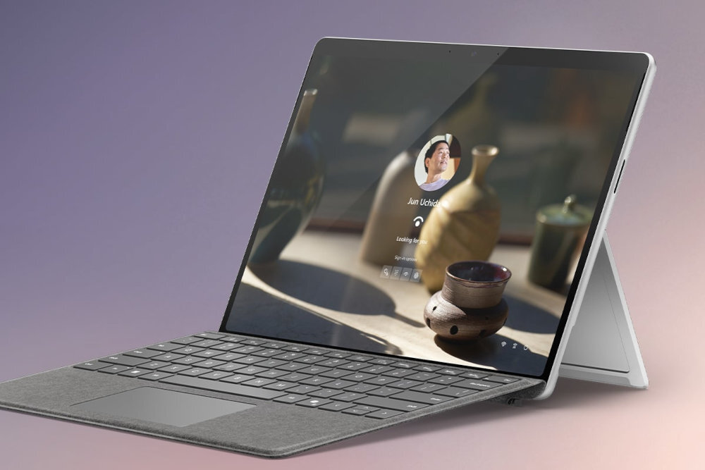Laptop z klawiaturą i ekranem dotykowym, na którym widoczny jest ekran logowania z awatarem i tekstem \'Jun Uchida\'. Urządzenie stoi na podstawce, a w tle widać rozmyte kształty naczyń.