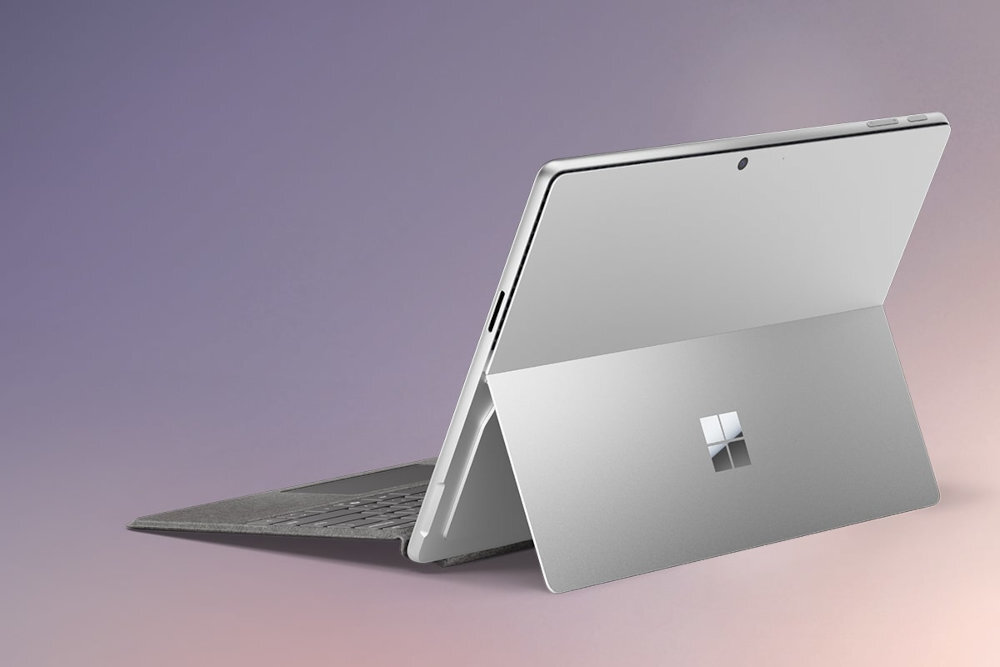 Na obrazie widoczny jest srebrny laptop z otwartą podpórką, na której znajduje się logo w formie czterech kwadratów. Tło jest gradientowe, przechodzące od fioletu do różu.