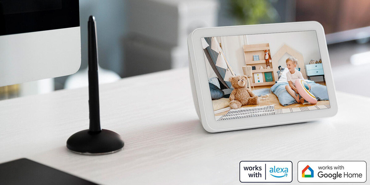 Na biurku stoi biały ekran wyświetlający obraz dziecka siedzącego na podłodze obok pluszowego misia. Obok ekranu znajdują się ikony z napisami: \'works with alexa\' i \'works with Google Home\'.