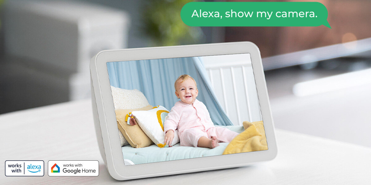 Na ekranie urządzenia widać uśmiechnięte dziecko siedzące na łóżku. W lewym dolnym rogu znajdują się ikony \'works with Alexa\' i \'works with Google Home\', a w prawym górnym rogu jest tekst w chmurce: \'Alexa, show my camera.\'
