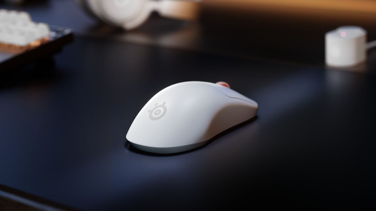 Na ciemnym biurku znajduje się biała mysz komputerowa z widocznym logo SteelSeries. W tle widać fragment klawiatury i niewielkie urządzenie elektroniczne.