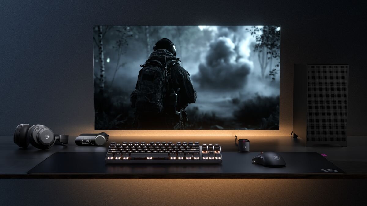 Na biurku znajduje się klawiatura, mysz z logo SteelSeries, słuchawki oraz monitor wyświetlający scenę z gry przedstawiającą postać w lesie. Obok monitora stoi głośnik, a całość jest oświetlona delikatnym światłem.