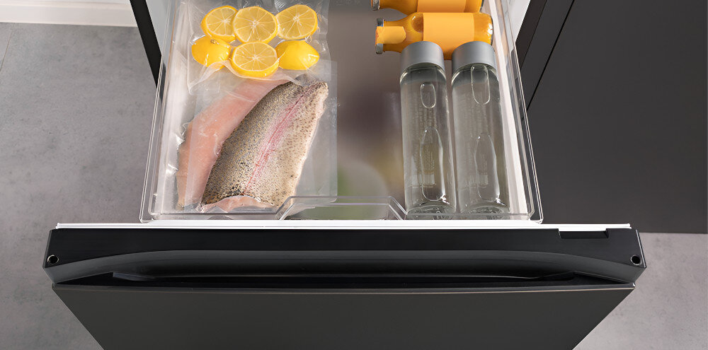 Otwarta szuflada lodówki z przechowywanymi produktami: plastry cytryny, kawałek ryby w folii, dwie butelki z przezroczystą cieczą i dwa żółte pojemniki. Szuflada ma przezroczyste ścianki, a lodówka jest w kolorze czarnym.