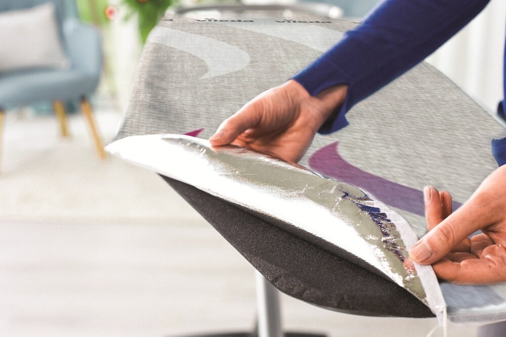Osoba trzyma pokrowiec deski do prasowania z metaliczną powierzchnią. W tle widoczne są rozmyte elementy wnętrza, takie jak krzesło i rośliny.